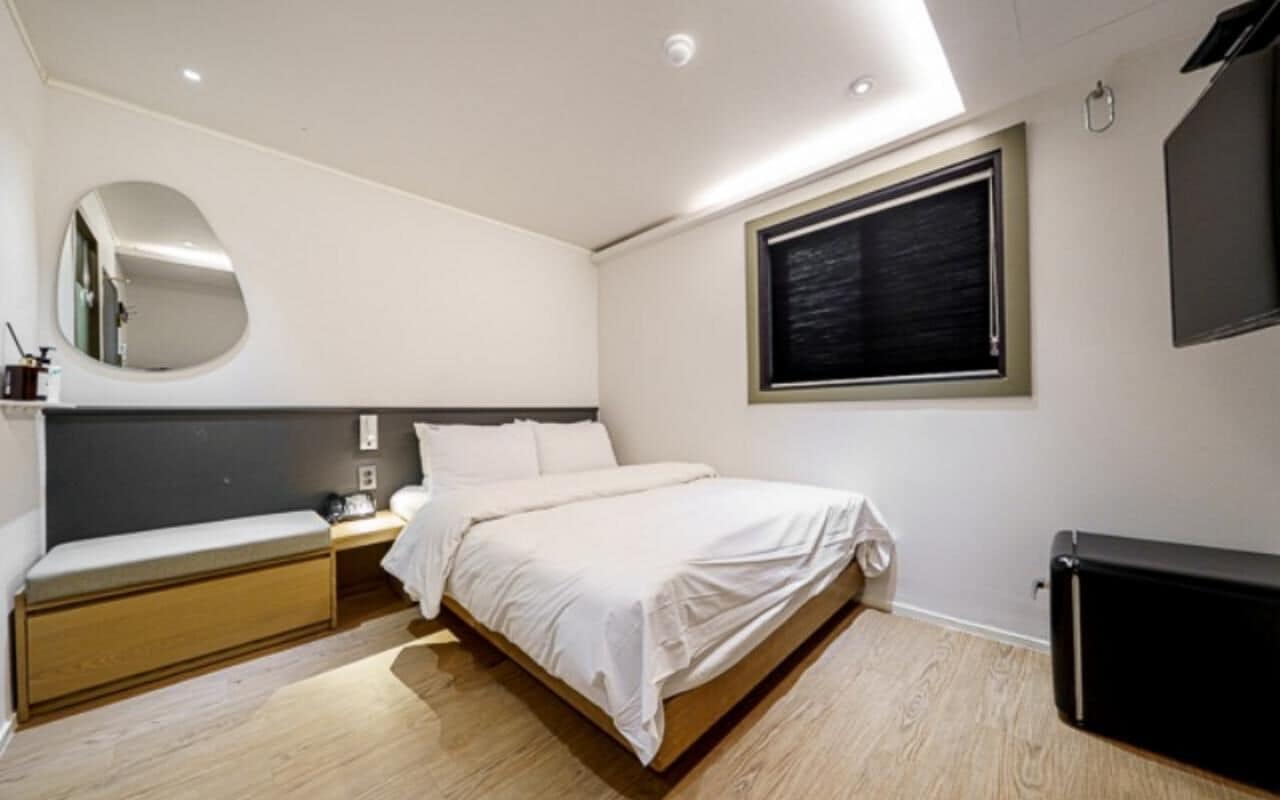Un dormitorio con cama, mesita de noche, banco y un ventanal grande.