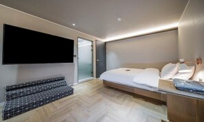 Habitación de hotel moderna con cama, televisor montado en la pared, zona de estar con sofá estampado y mesita de noche de madera.