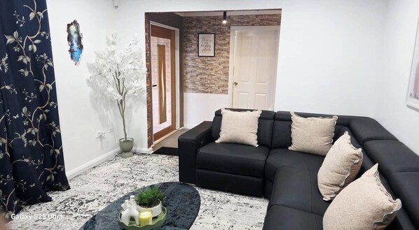 Un salon avec un canapé noir, des coussins décoratifs, une petite table sur laquelle est posée une plante, et un tableau accroché au mur.