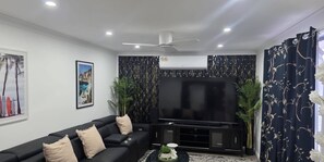 Un salon avec un canapé noir, un téléviseur à écran plat, des panneaux décoratifs sur les murs et un ventilateur de plafond.
