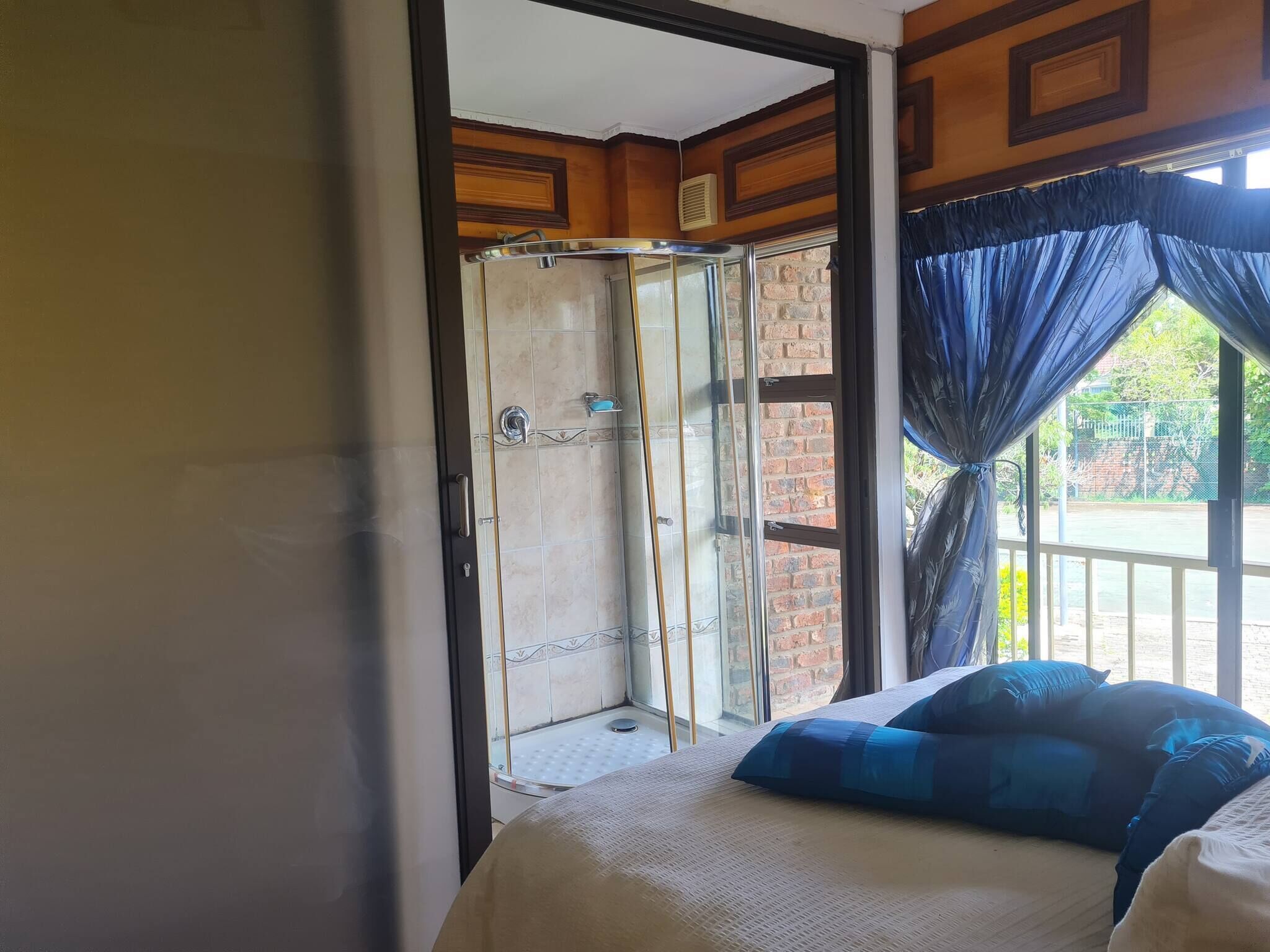Una habitación con cama, ducha con puertas de cristal, ventana con cortinas y vista a un muro de ladrillo y follaje exterior.