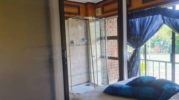 Una habitación con cama, ducha con puertas de cristal, ventana con cortinas y vista a un muro de ladrillo y follaje exterior.