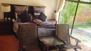 A bedroom with a bed, two upholstered chairs, a nightstand, and a window with a view of greenery.