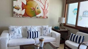 Ein Wohnzimmer mit einem weißen Sofa, gestreiften Kissen, einem Bild von Fischen, einer Lampe und einem Fenster.