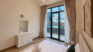 Ein modernes Schlafzimmer mit einem großen Bett, einer Kommode, einem Spiegel und Blick auf die Stadt durch ein großes Fenster.