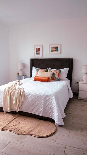 A neatly made bed with a white quilt, a brown throw blanket, and a variety of pillows. There are two bedside tables with lamps, a wall-mounted picture frame, and a wooden floor.
