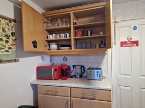 Une petite cuisine équipée d’un four à micro-ondes, d’une machine à café et d’un grille-pain sur le plan de travail, d’un meuble en bois au-dessus et d’un panneau « Il est interdit de fumer » sur la porte.