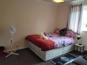Une chambre à coucher avec un lit, un ventilateur sur pied, une petite table et une fenêtre avec des rideaux.