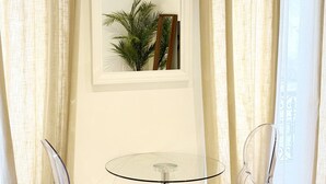 A room with a round glass table, two clear chairs, a potted plant, and beige curtains. - Charming studio in the heart of République (Paris)