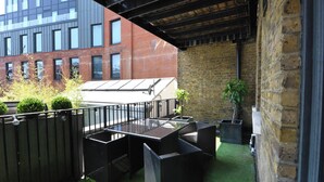 Un patio al aire libre con una mesa de vidrio y mobiliario de mimbre negro, una pared de ladrillo y un edificio moderno al fondo.