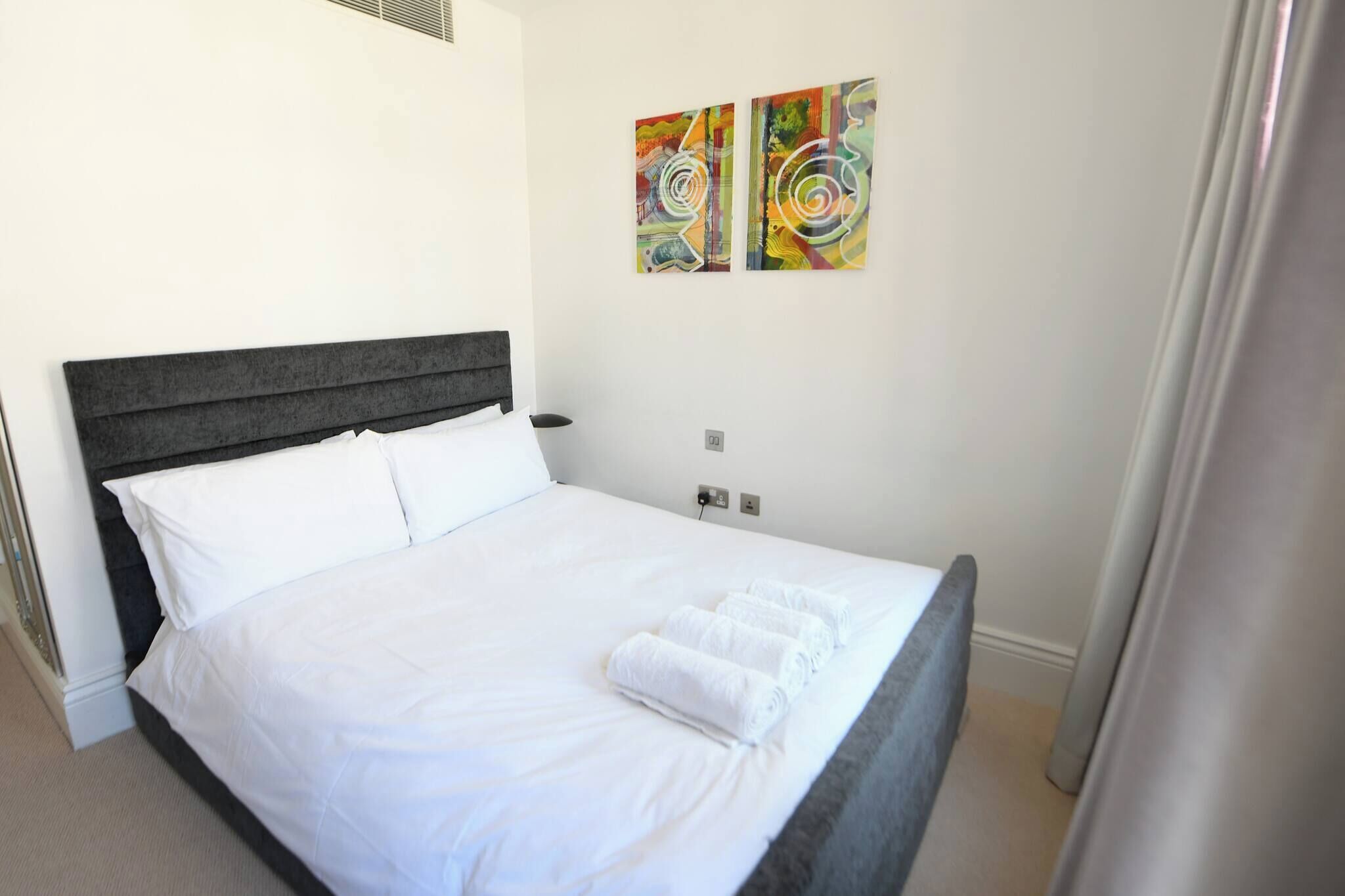 A neatly made bed with white linens and rolled towels, a dark headboard, and two abstract paintings on the wall.