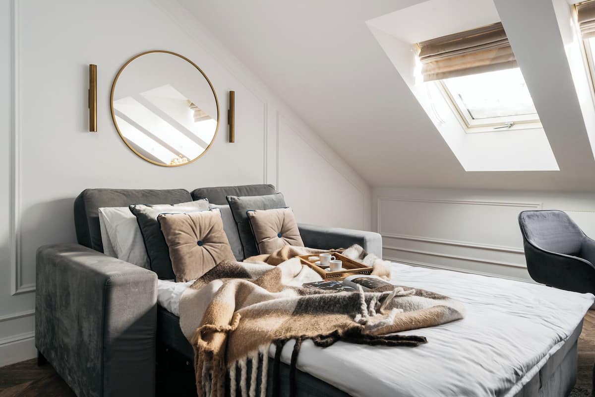 A modern bedroom with a loft-style ceiling, a large bed with multiple pillows, a side table, and a window with blinds.