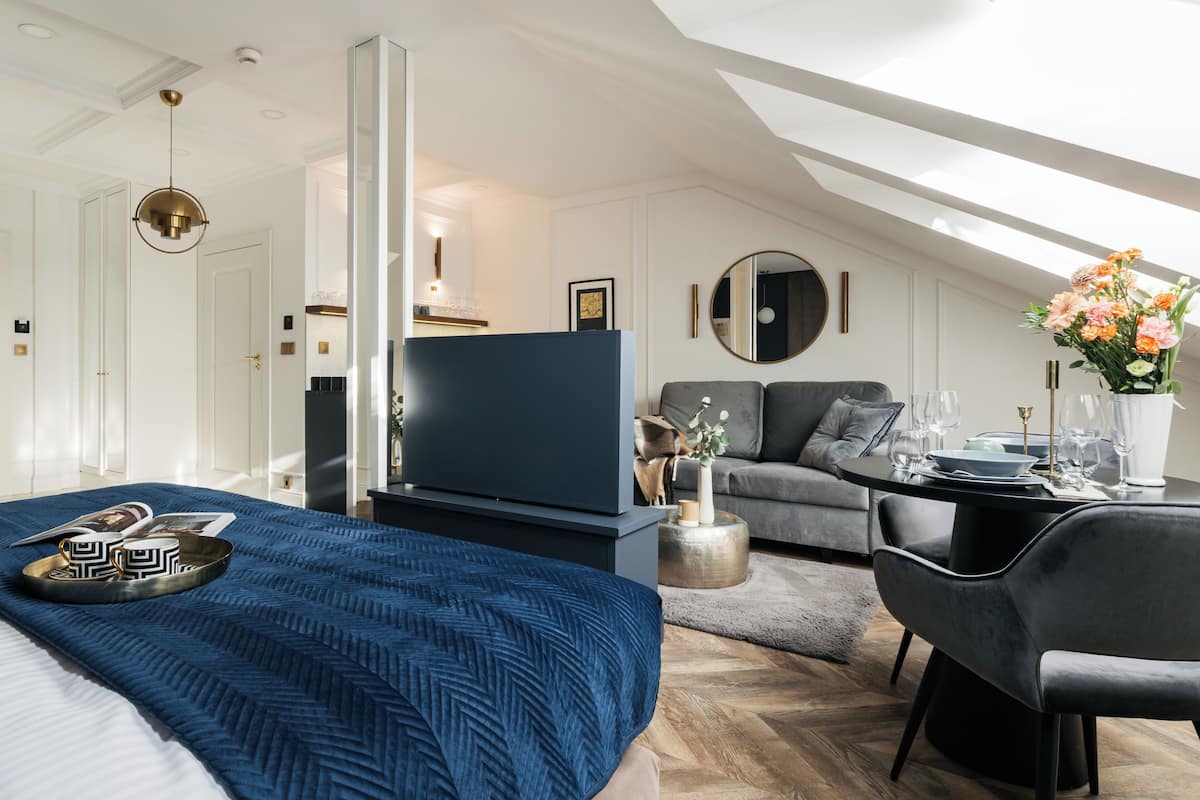 A modern hotel room with a large bed, a flat-screen TV, a sofa, a dining table with chairs, and a decorative vase with flowers.