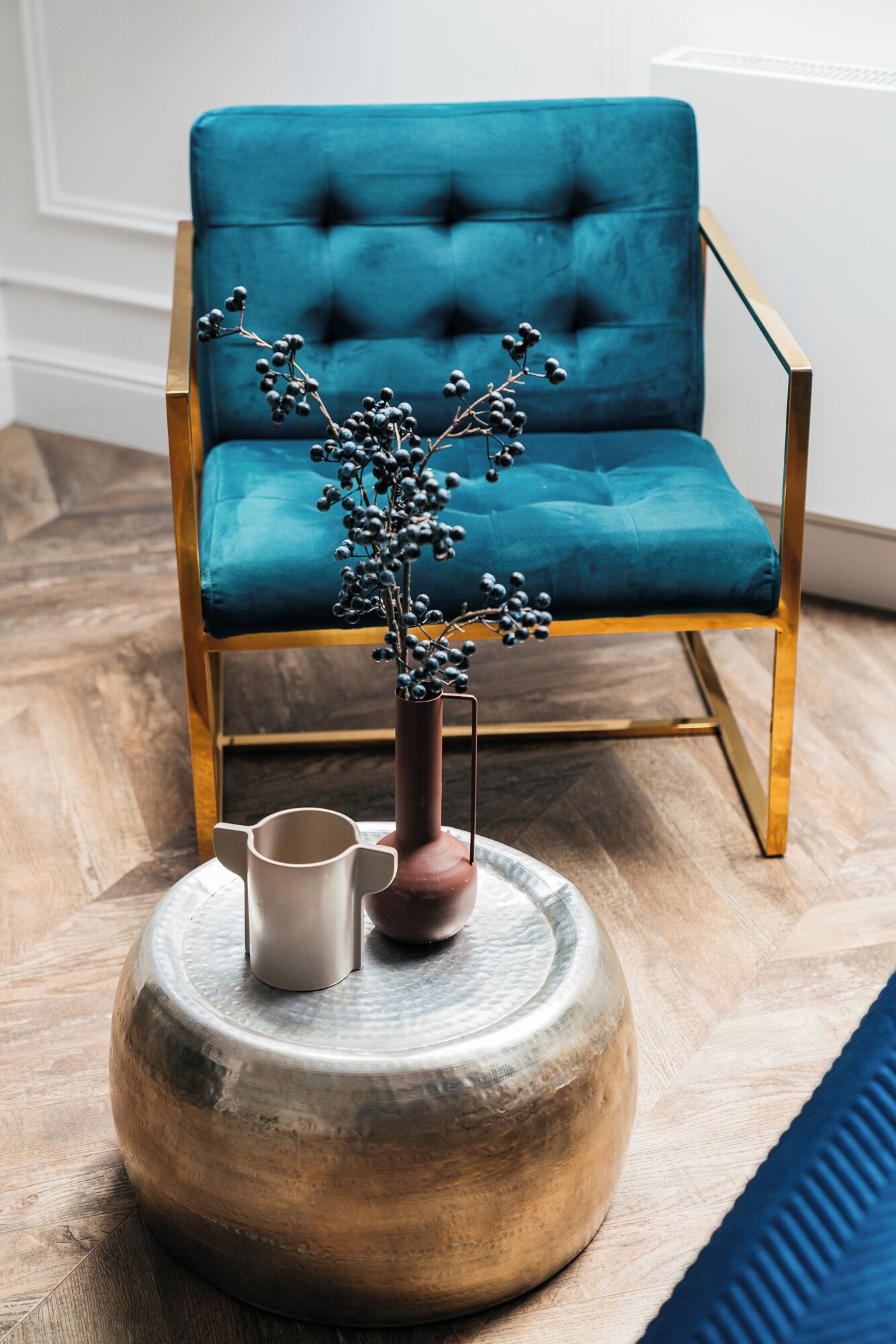 A teal upholstered chair with a gold frame, a round wooden table with a vase and a mug, and a blue cushion on the floor.