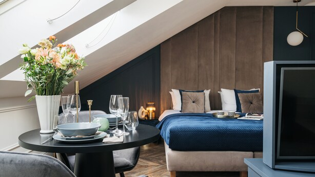 A modern hotel room with a bed, a TV, a dining table with a vase of flowers, and a view of the sky through a skylight. - Central Palace (Kraków)