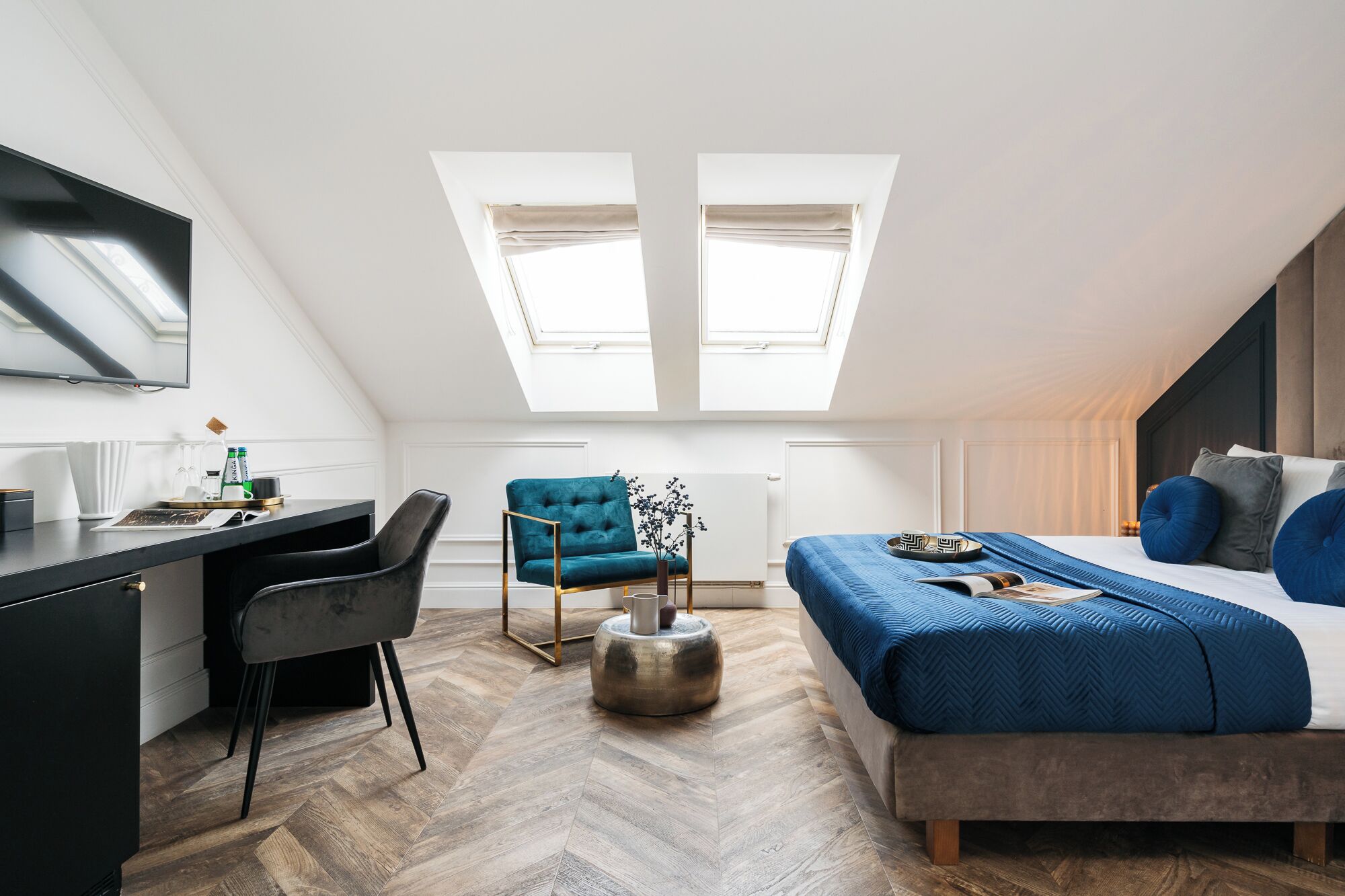 A modern loft bedroom with a desk, a blue sofa, a bed with blue bedding, and two skylights.