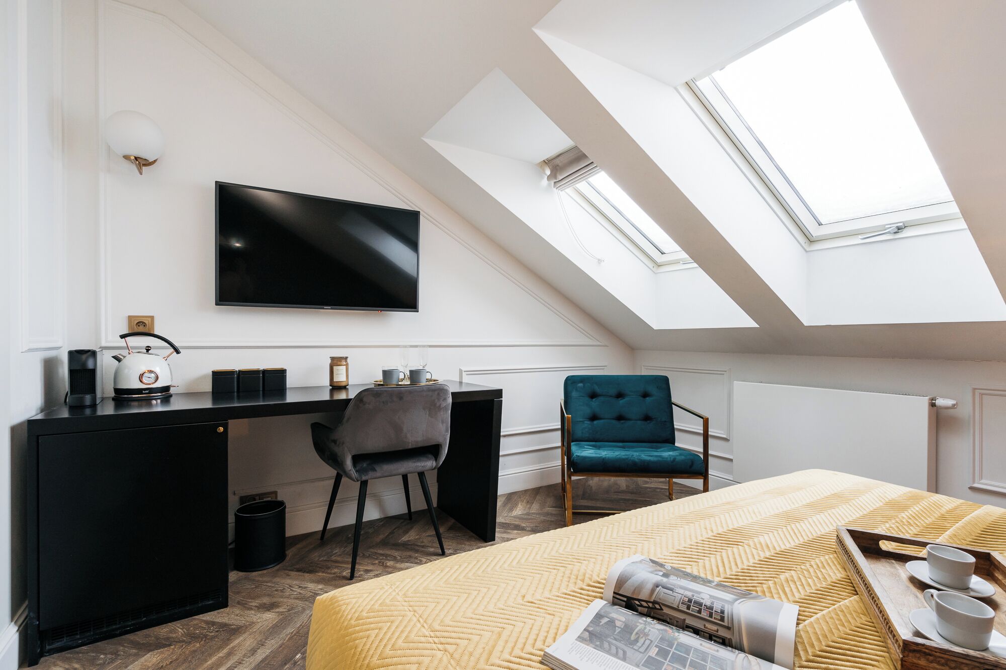 A modern room with a skylight, a flat-screen TV, a desk, a chair, and a bed with a magazine and cups.