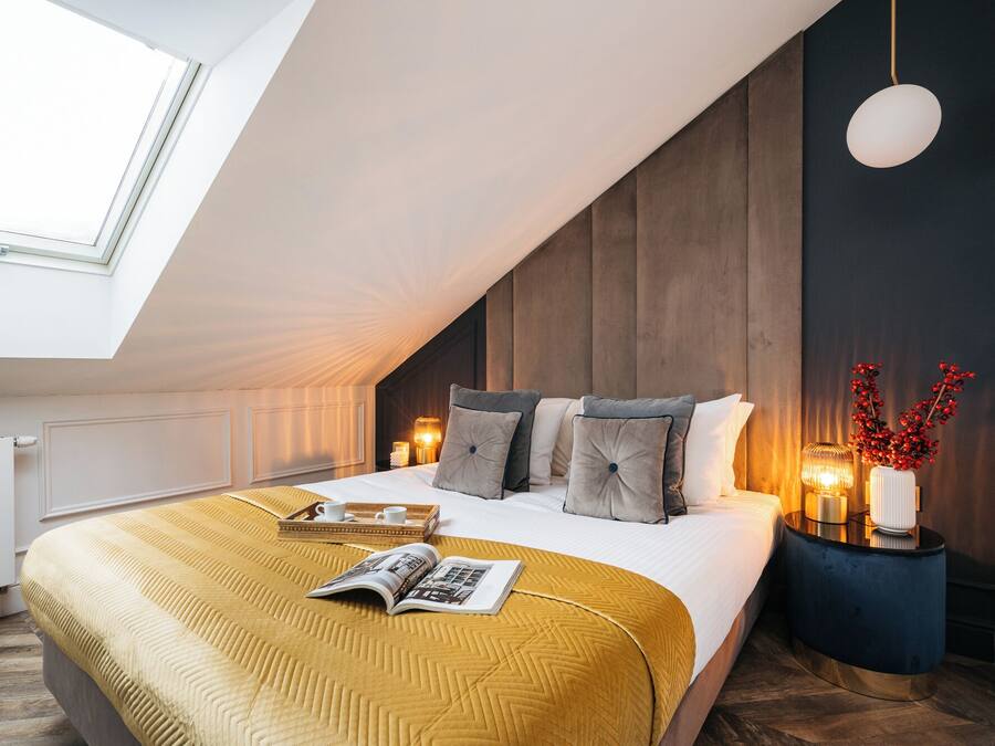 A modern bedroom with a sloped ceiling, a large bed with a mustard-colored blanket, bedside lamps, and a decorative vase with red berries.