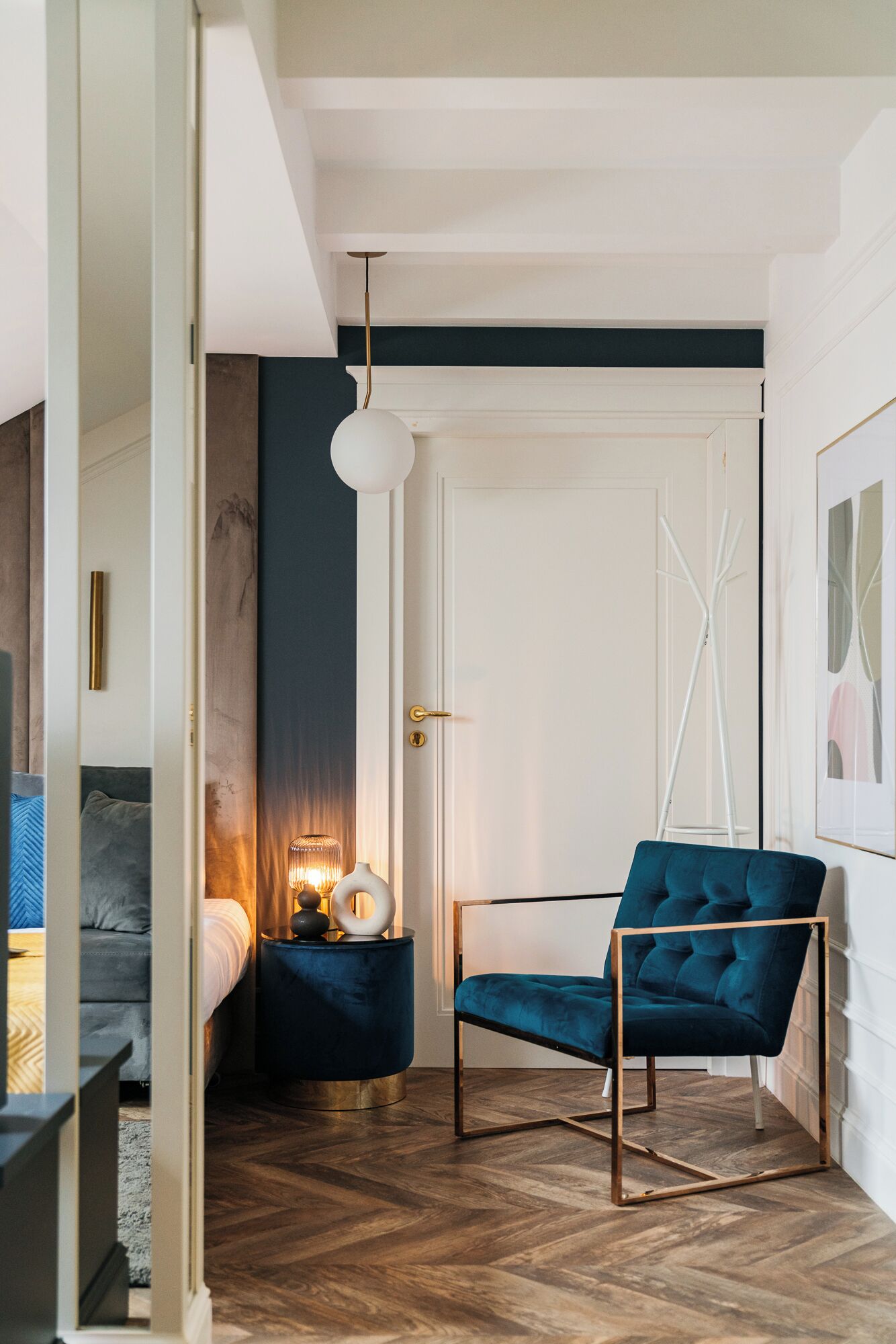 A modern bedroom with a bed, a blue chair, a bedside table with a lamp, and a door with a white frame.