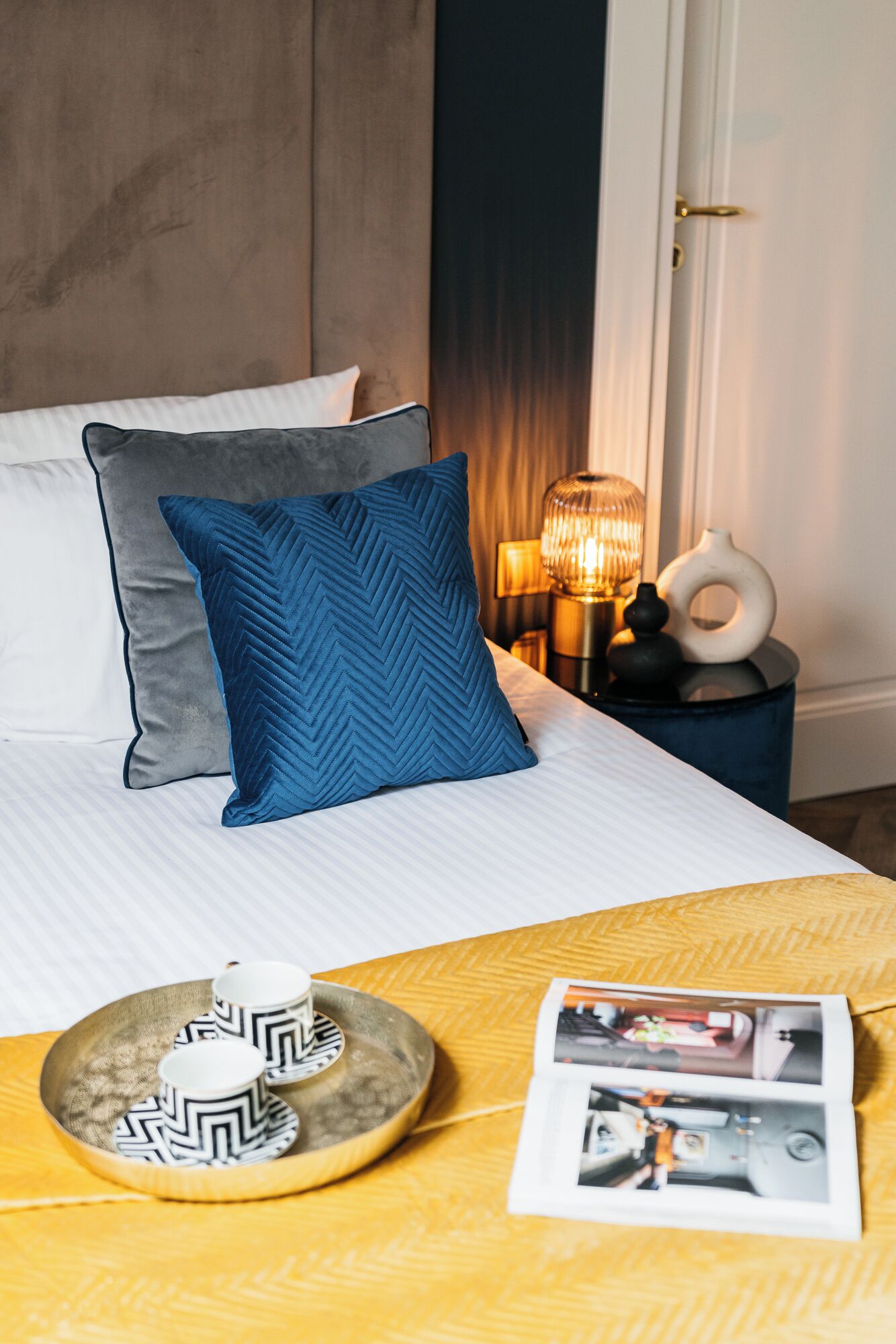 Una cama bien hecha con almohadas, una mesita de noche con lámpara, y una bandeja con tazas y platillos.