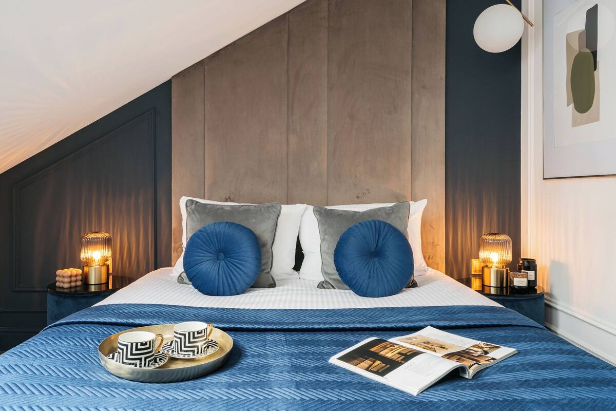 A bedroom with a sloped ceiling, a bed with a blue quilt, two bedside lamps, and a magazine on the bed.