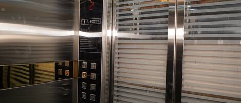 An elevator interior with metallic walls, a control panel with buttons, and horizontal blinds.