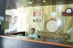 A reception desk with a clock, a framed picture, and a sign indicating check-in and check-out times. - Hotel Vedant (Jaipur)
