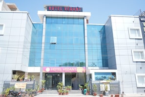 A modern hotel with a large glass facade, a signboard with 'HOTEL VEDANT' and 'ACT LUXURY ROOM' text, and potted plants at the entrance. - Hotel Vedant (Jaipur)