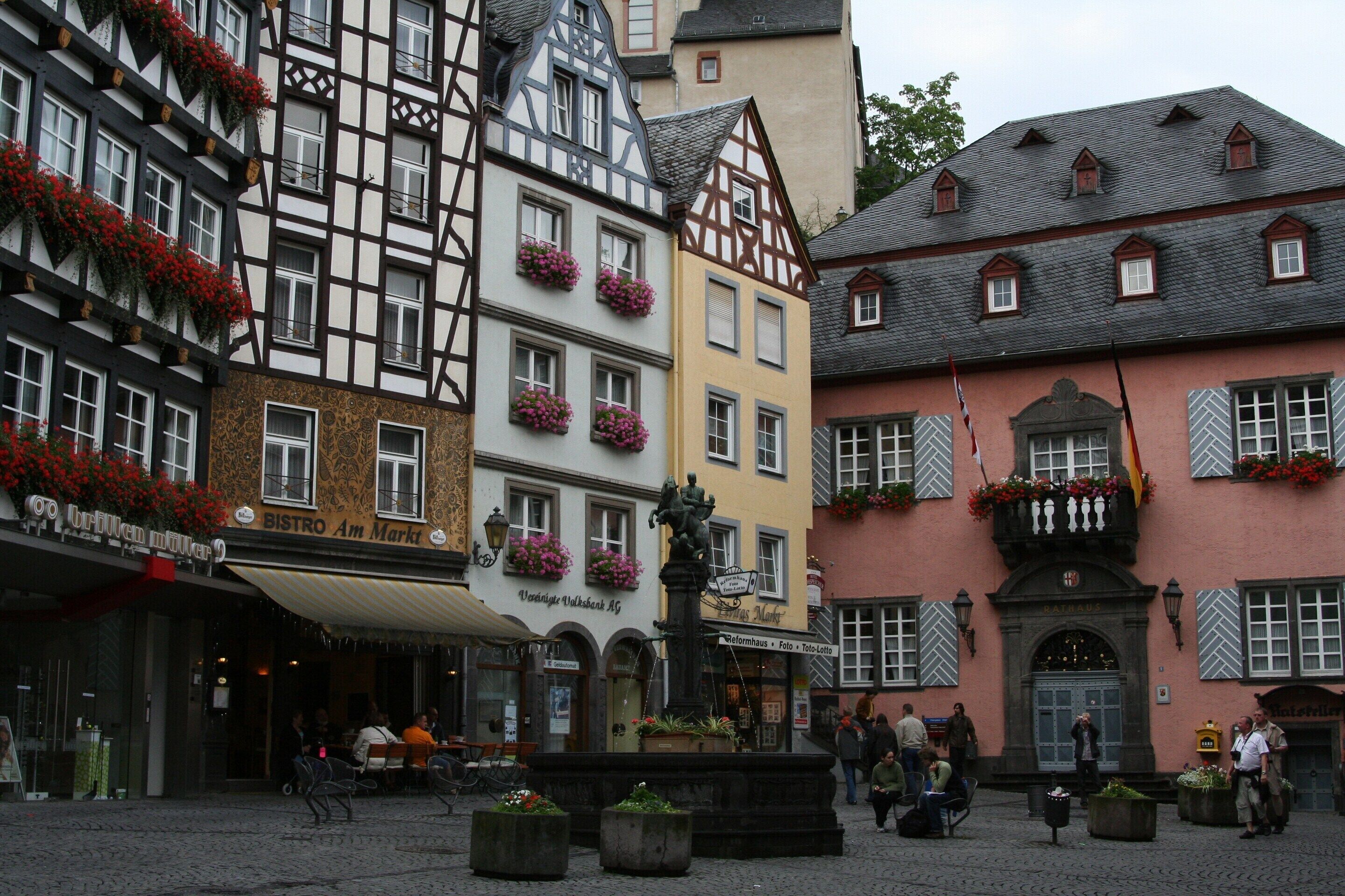 A European street with traditional half-timbered buildings, a fountain, and outdoor seating areas.