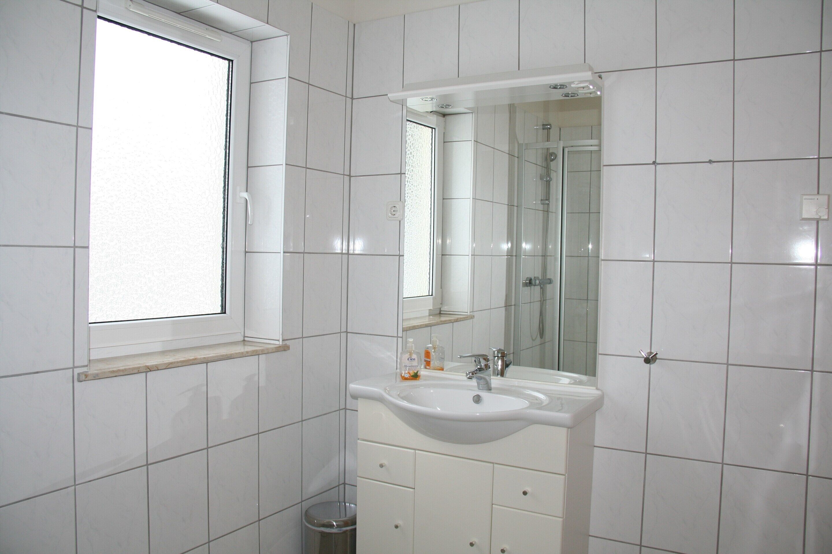 Ein Badezimmer mit weißen Fliesen, einem Fenster, einem Waschbecken und einem Spiegel.
