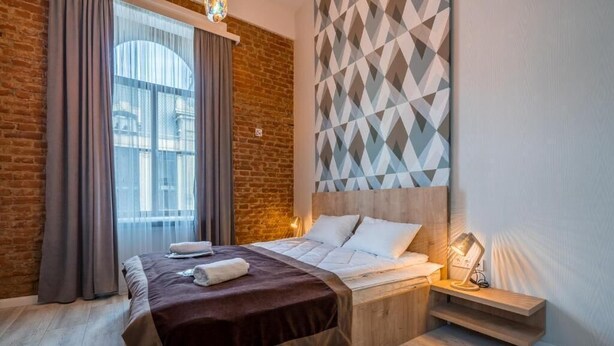 Ein Schlafzimmer mit Backsteinwand, einem geometrisch gemusterten Kopfteil, einem Bett mit weißen Bettwäsche, einem Nachttisch mit Lampe und einem Fenster mit Vorhängen.