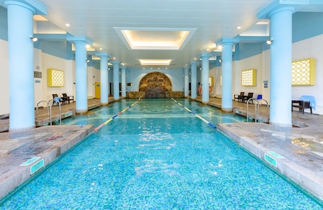 A large indoor swimming pool with clear water, surrounded by columns and benches, and a stone wall at the far end.