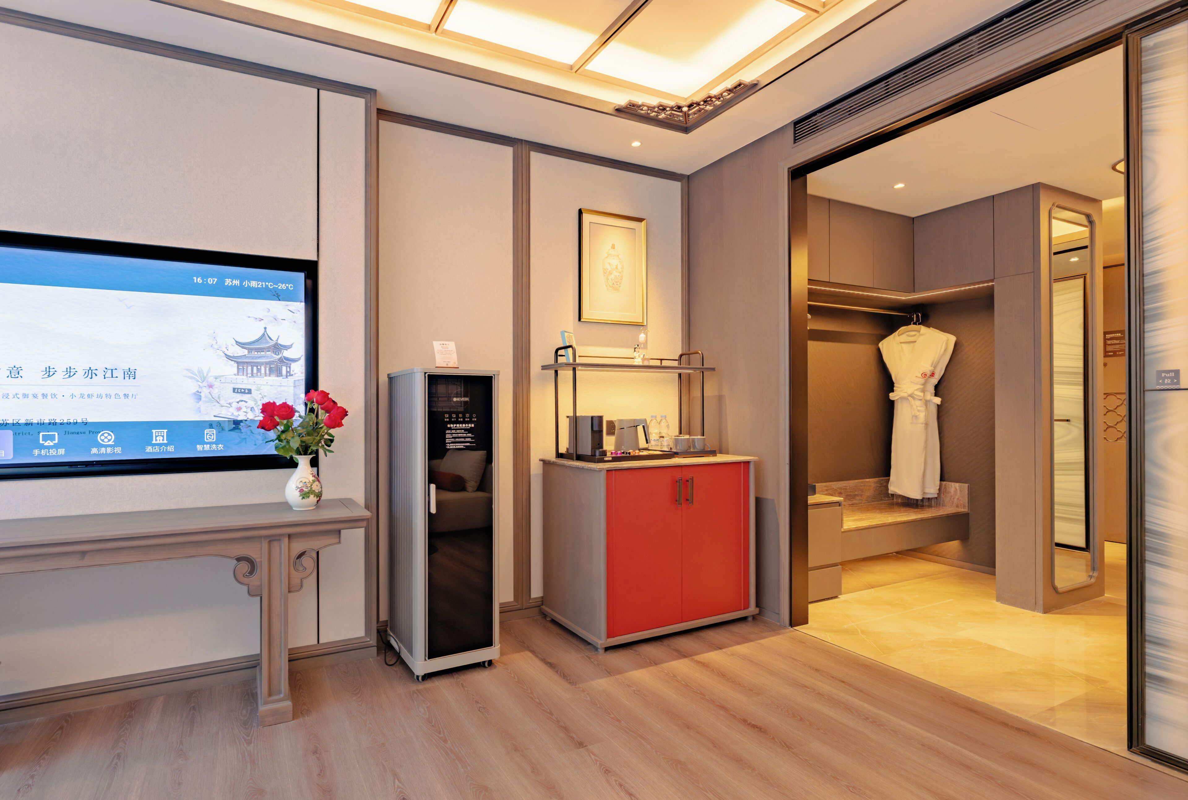 A hotel room with a television displaying a weather forecast, a small table with a vase of flowers, a red cabinet, and a bathroom with a robe hanging.