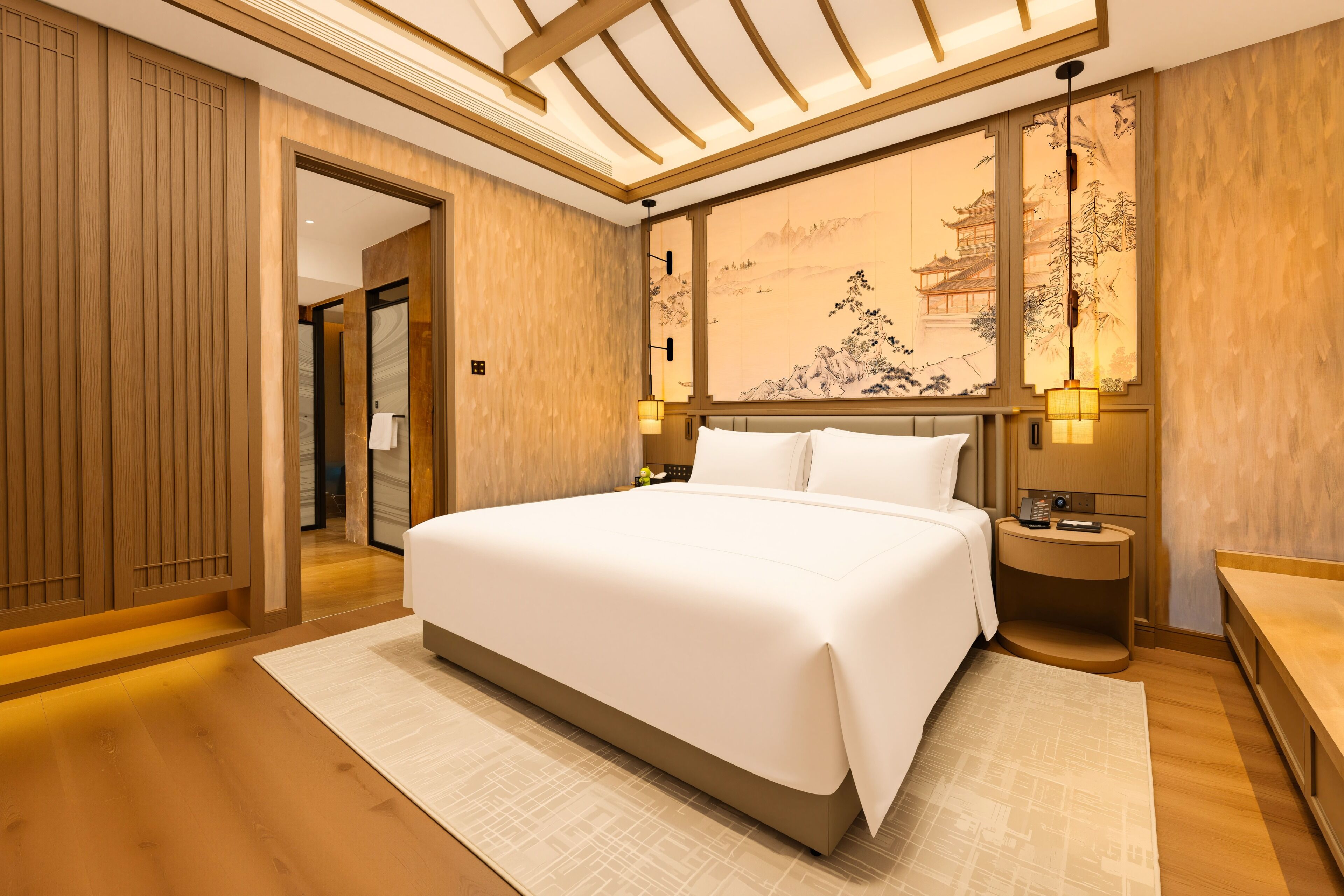 Quarto de hotel moderno com uma cama grande, painéis de madeira, uma obra de arte tradicional na parede e uma pequena mesa de cabeceira com abajur.
