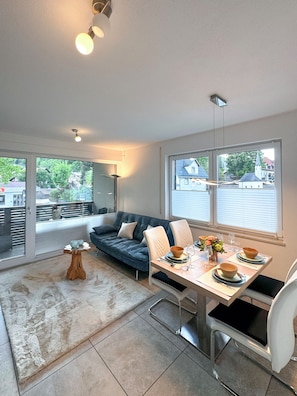 A modern living room with a sofa, dining table, and chairs. There is a large window with a view of the outside.
