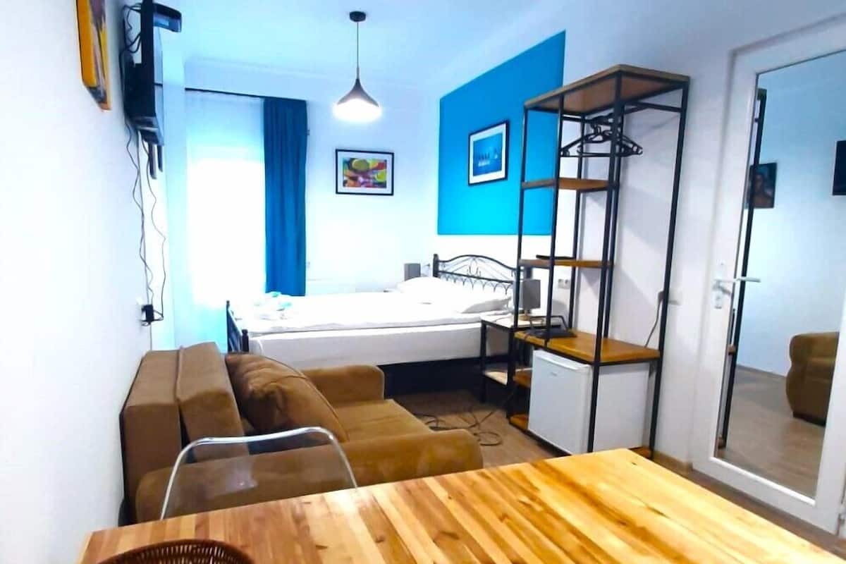 A room with a bed, a wooden table, a sofa, a small desk, a hanging light fixture, and a wall-mounted shelf.