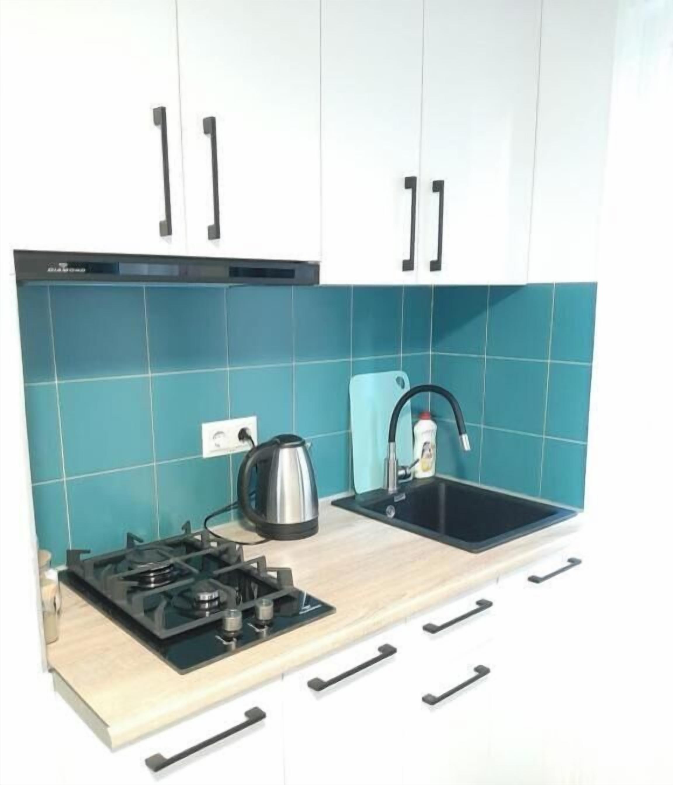 A kitchen with teal tiles, a gas stove, a kettle, and a sink.