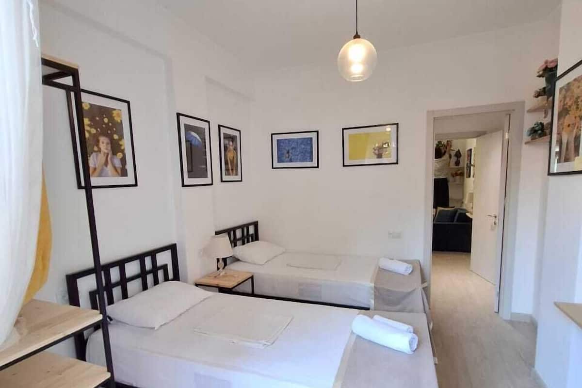 A room with two beds, a wooden desk, a hanging light fixture, and framed pictures on the wall.