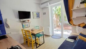 Un salon moderne comprenant un canapé, une table à manger avec des chaises, un téléviseur fixé au mur et une porte-fenêtre coulissante donnant sur un espace extérieur.