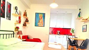 Une chambre avec un lit, un canapé rouge, un bureau avec deux chaises, un coin cuisine avec des placards blancs et un tableau au mur.