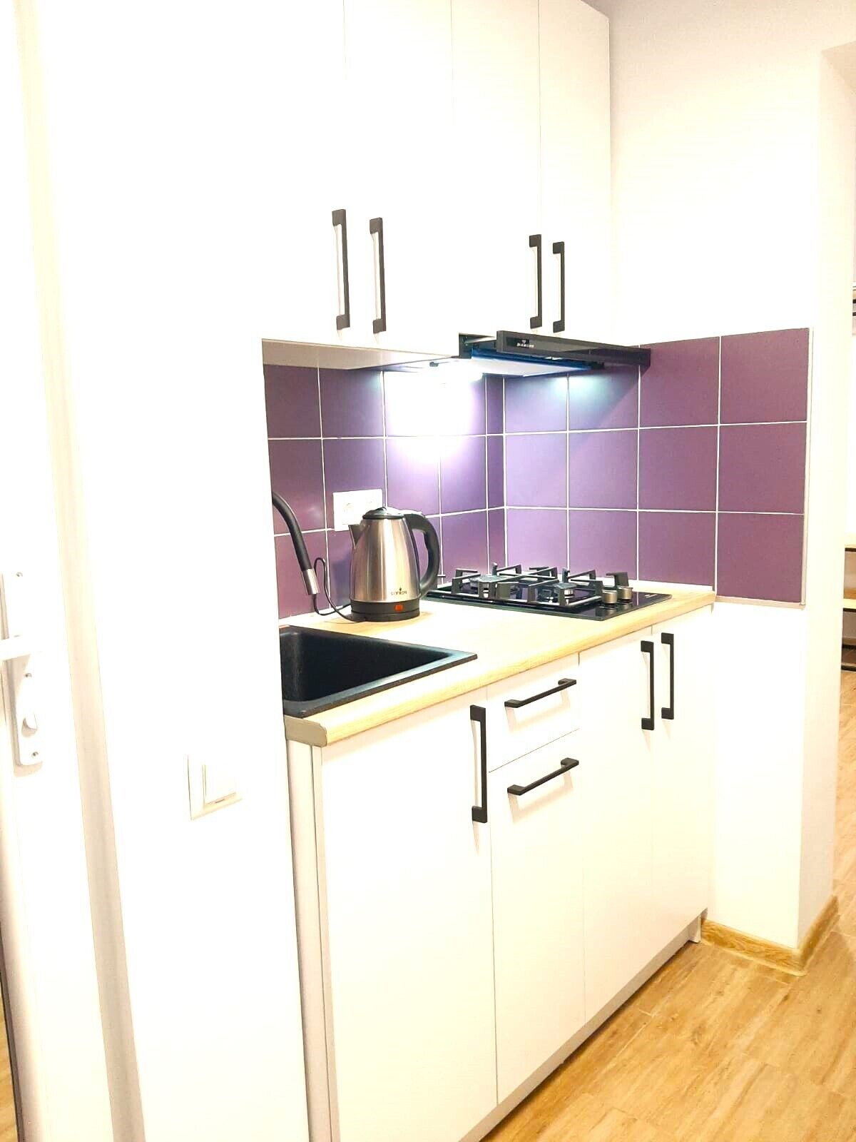 A compact kitchen with white cabinets, a gas stove, a kettle, and purple-tiled backsplash.