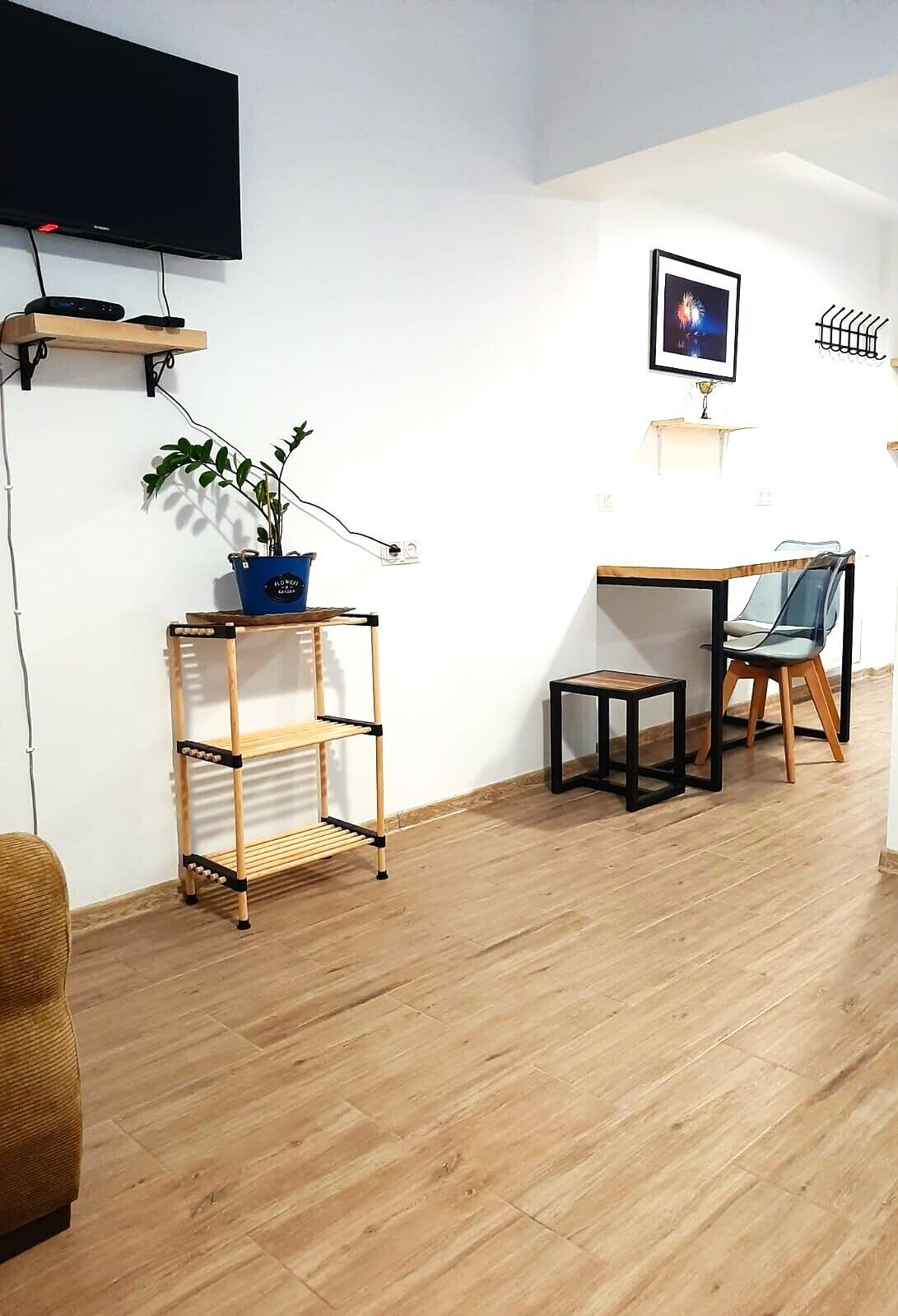 A room with a flat-screen TV, a wooden floor, a small table with a chair, a plant on a shelf, and a framed picture on the wall.