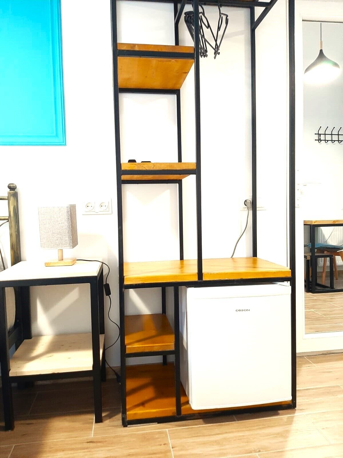 Quarto compacto com um mini-frigorífico branco, uma escrivaninha de madeira com uma luminária e uma parede azul.