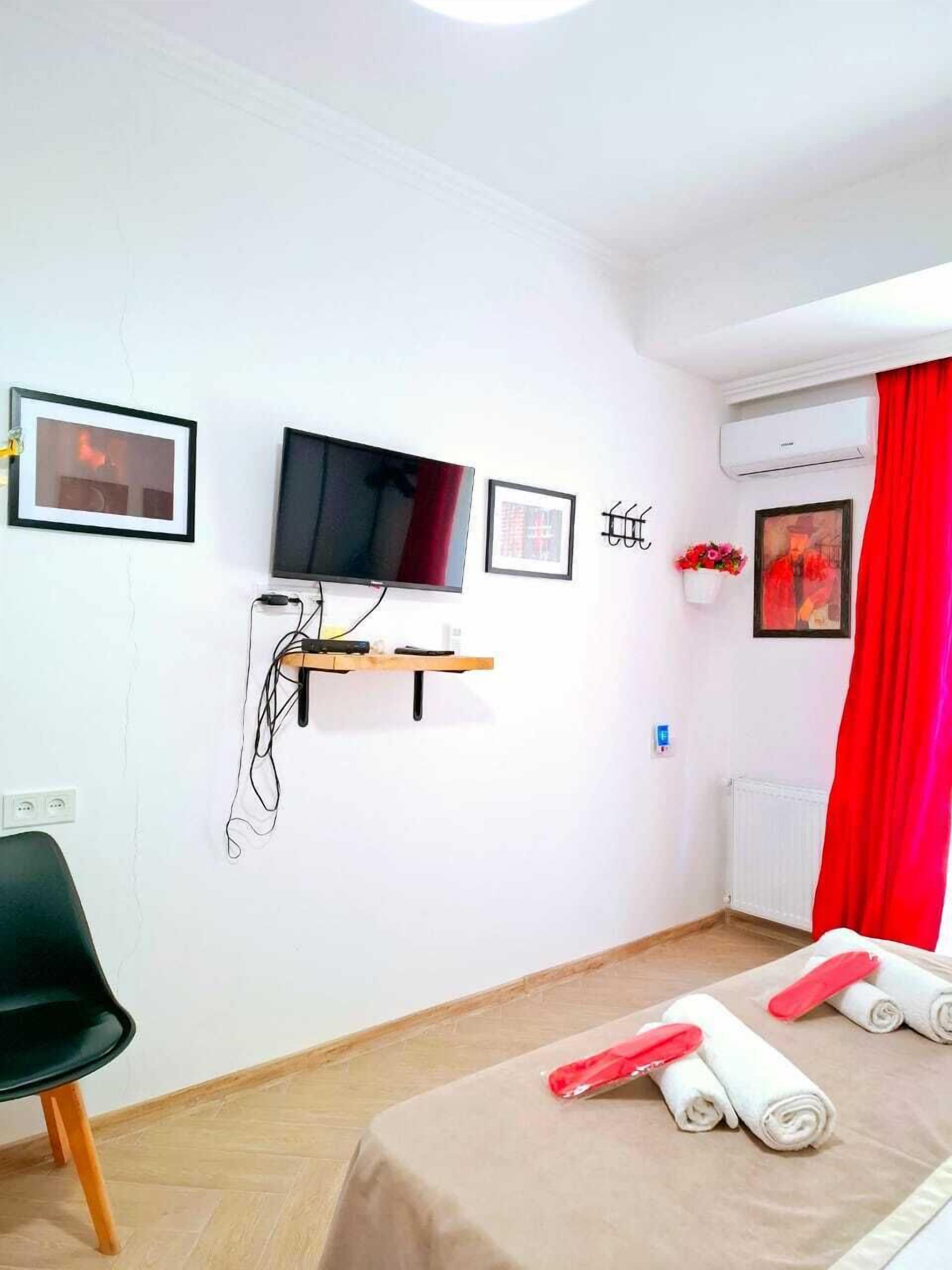 A hotel room with a bed, a chair, a TV mounted on the wall, a radiator, and a window with red curtains.