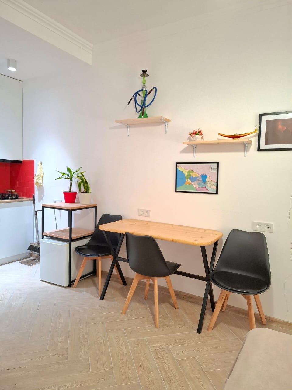 Een ruimtelijk ingerichte leefruimte met een houten eettafel, zwarte stoelen, een kleine keukenhoek, een potplant en wandplanken met een kaart en foto.