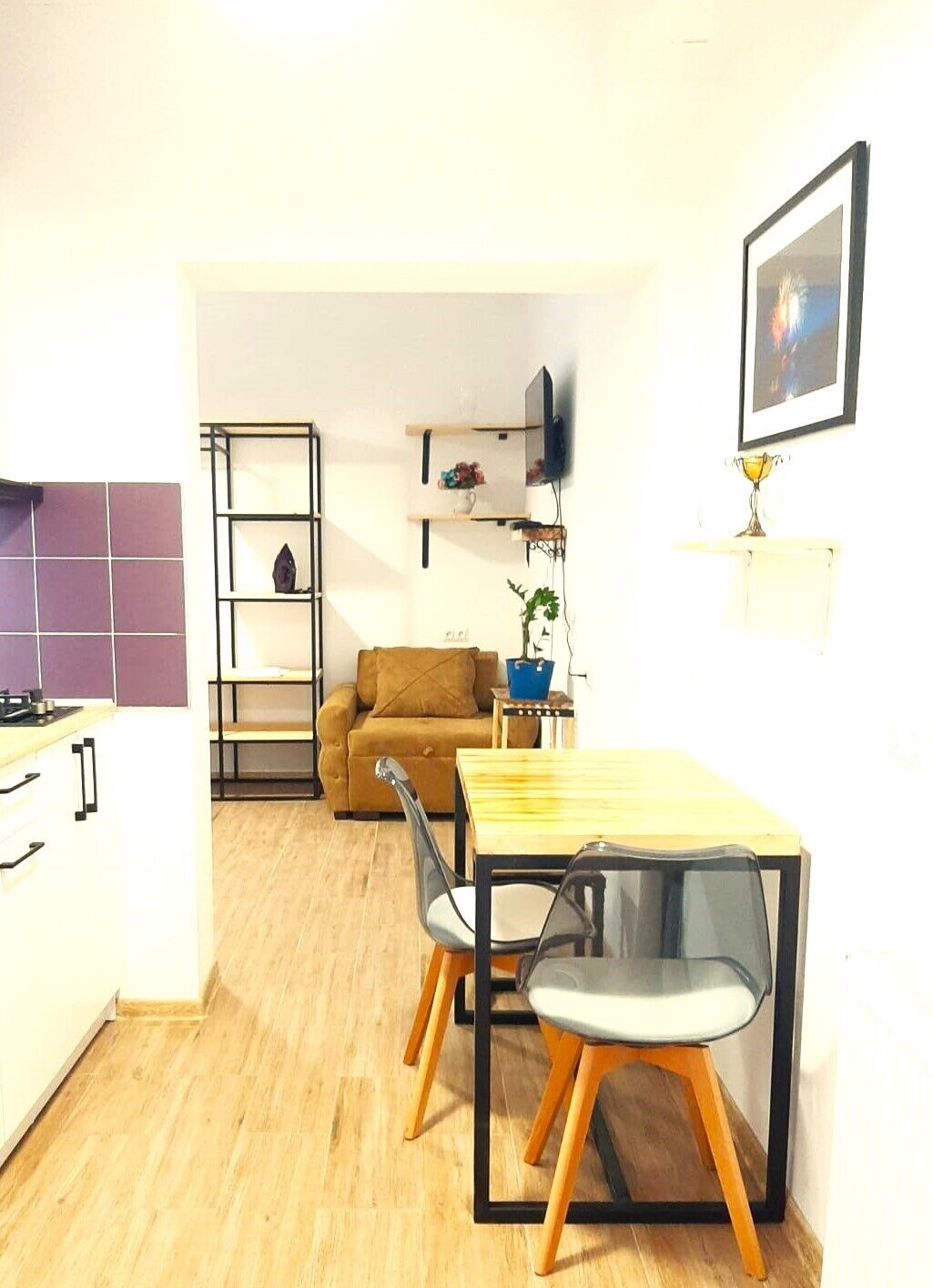 Een compacte leefruimte met een kitchenette, een eethoek met twee stoelen, een bank en een aan de muur bevestigde televisie.