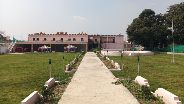 A pathway leading to a building with a pink facade and arched windows, surrounded by a green lawn and a few tents. - Kaku's Krishnabagh palace (Raipur Karchuliyan)
