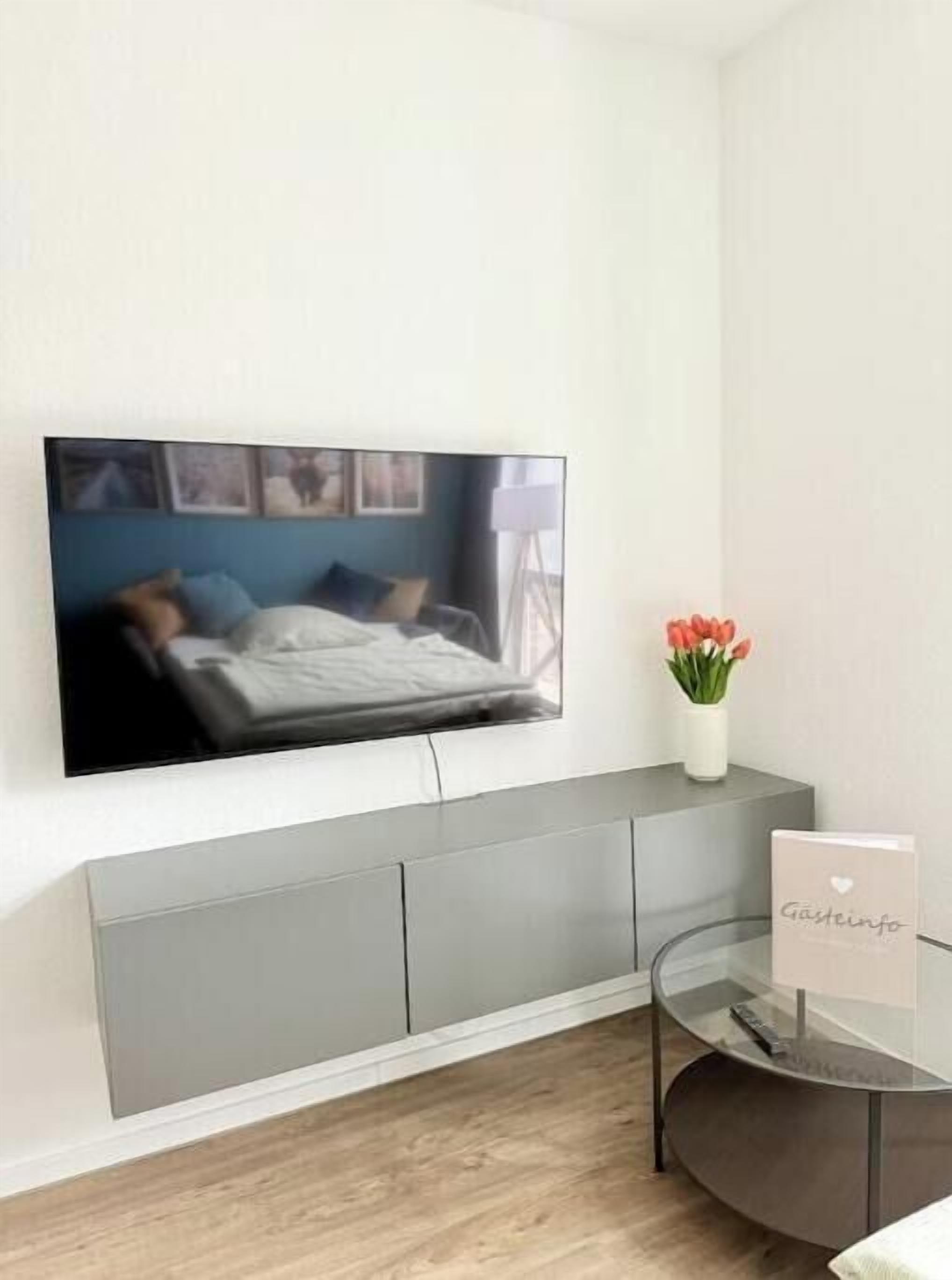Una sala moderna con un televisor de pantalla plana montado en la pared, una mesita consola gris y una pequeña mesa de vidrio con un letrero.