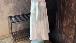 Un peignoir blanc est drapé sur un mannequin dans une pièce dont le sol est en bois, un mur est recouvert de carreaux et un panneau en bois sombre.