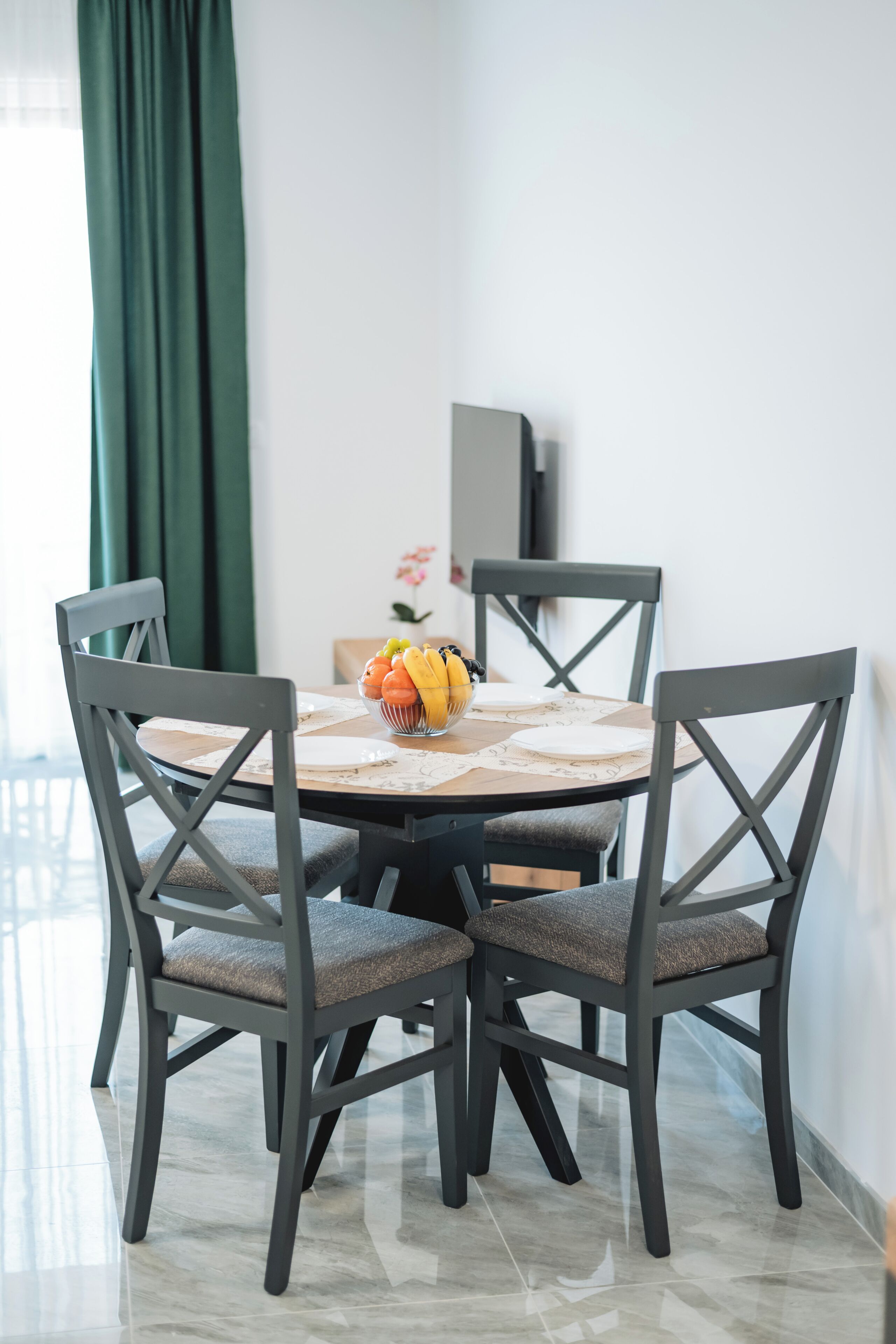 Un comedor con una mesa redonda, cuatro sillas, un frutero y una cortina verde.